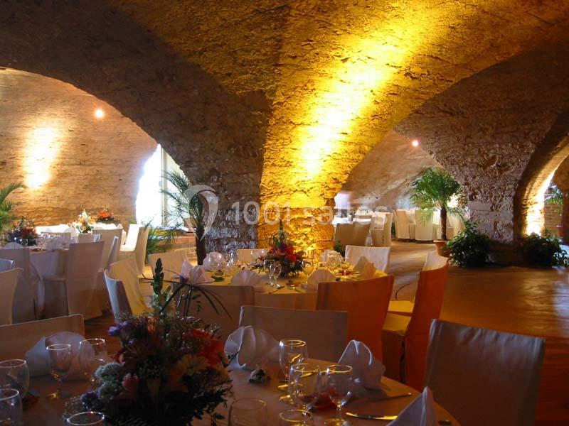 Salle voûtée en pierre éclairée, aménagée avec des tables décorées pour un événement ou un repas festif.