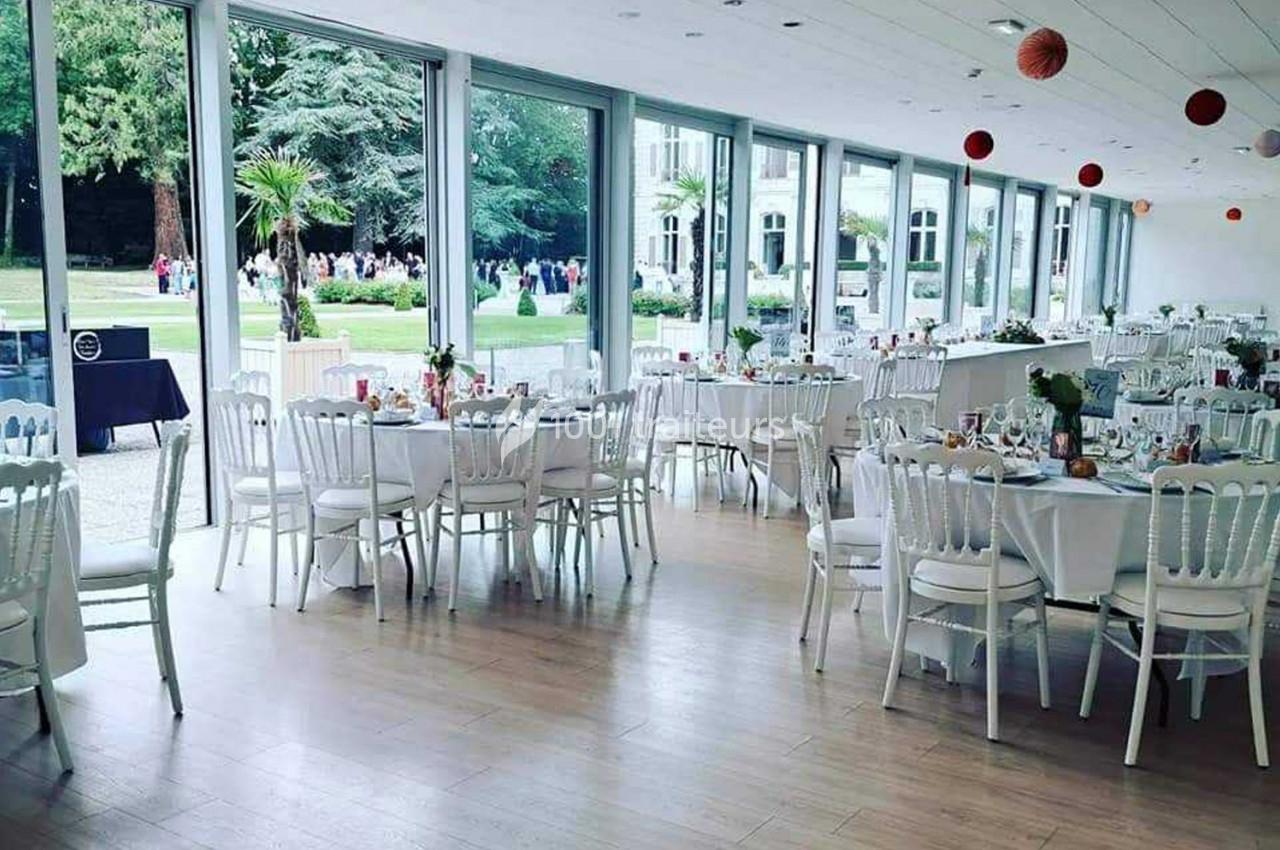 Salle lumineuse avec grandes baies vitrées, tables rondes dressées pour un événement, vue sur un jardin extérieur.