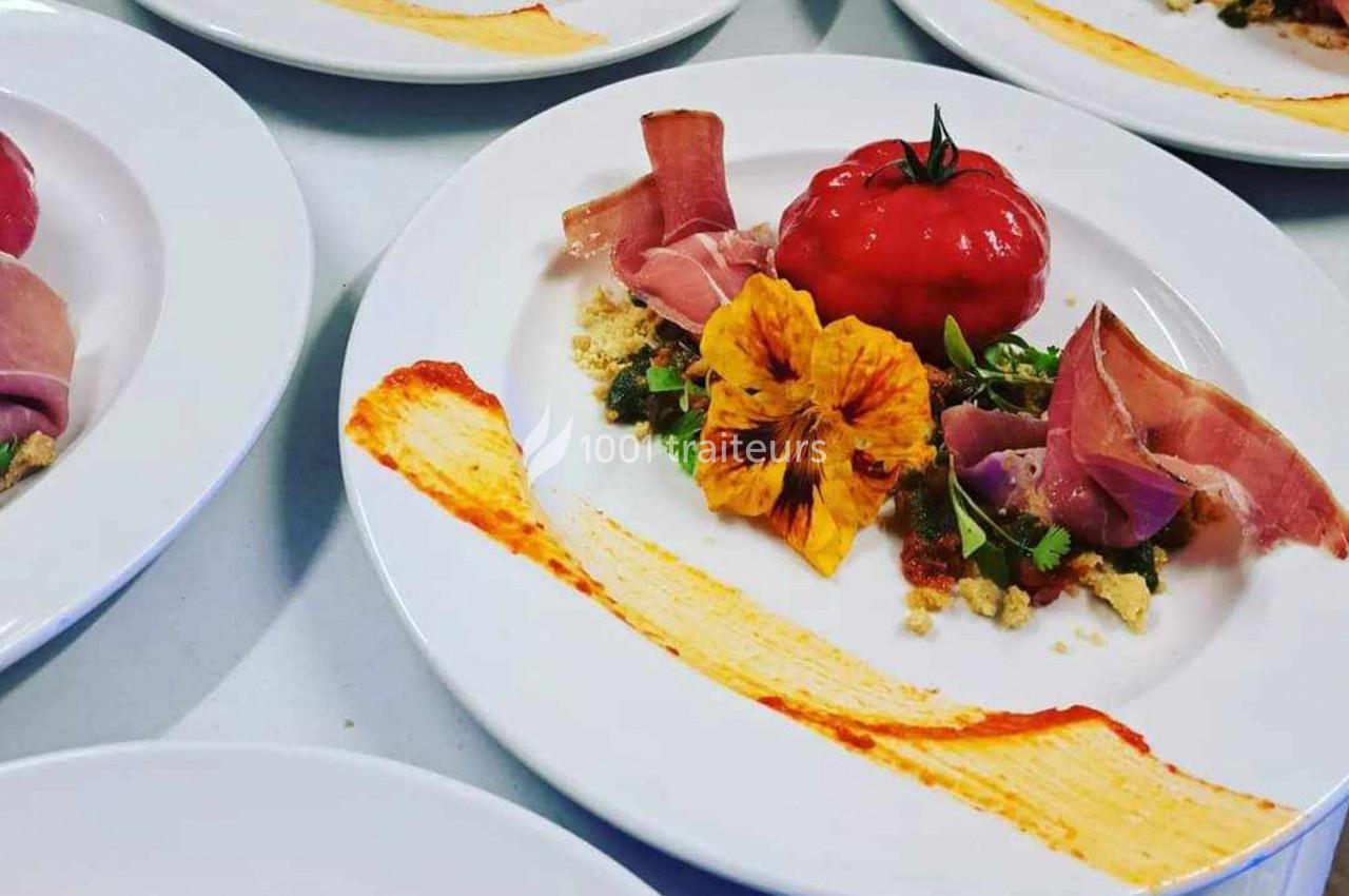 Assiette gastronomique avec tomate entière, jambon, fleur comestible et sauce colorée disposée artistiquement.