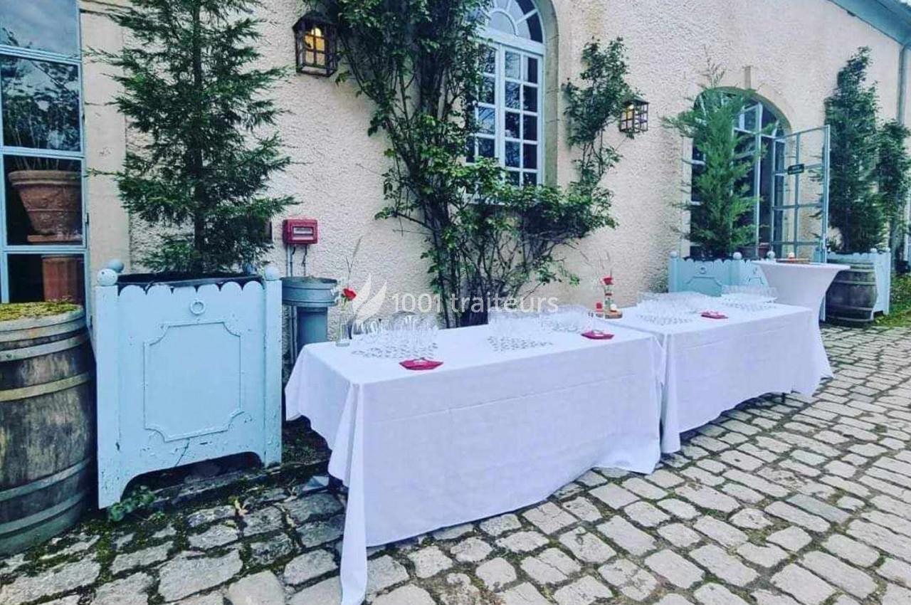 Tables dressées avec nappes blanches, verres et décorations, devant un bâtiment en pierre avec fenêtres et végétation.