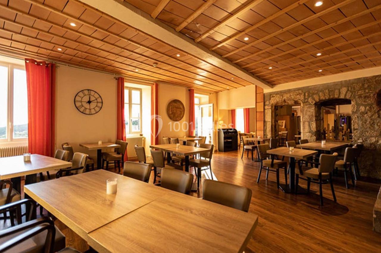 Salle de restaurant lumineuse avec tables en bois, chaises modernes, murs en pierre et grandes fenêtres avec rideaux rouges.
