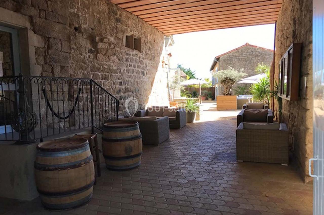 Passage couvert en pierre avec des tonneaux, des fauteuils en osier et une vue sur une cour ensoleillée avec des plantes.