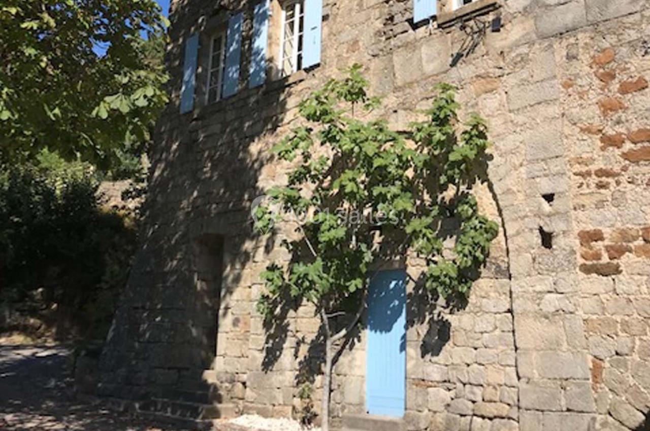Façade en pierre d'une maison avec volets bleus, porte bleue et un figuier devant, éclairée par le soleil.