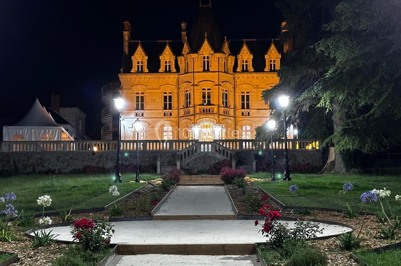 Façade illuminée d'un château de style classique, vue de nuit avec un jardin aménagé et des lampadaires au premier plan.