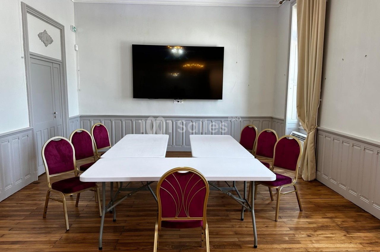 Salle lumineuse avec parquet, table blanche rectangulaire et chaises rouges disposées autour, écran mural en arrière-plan.