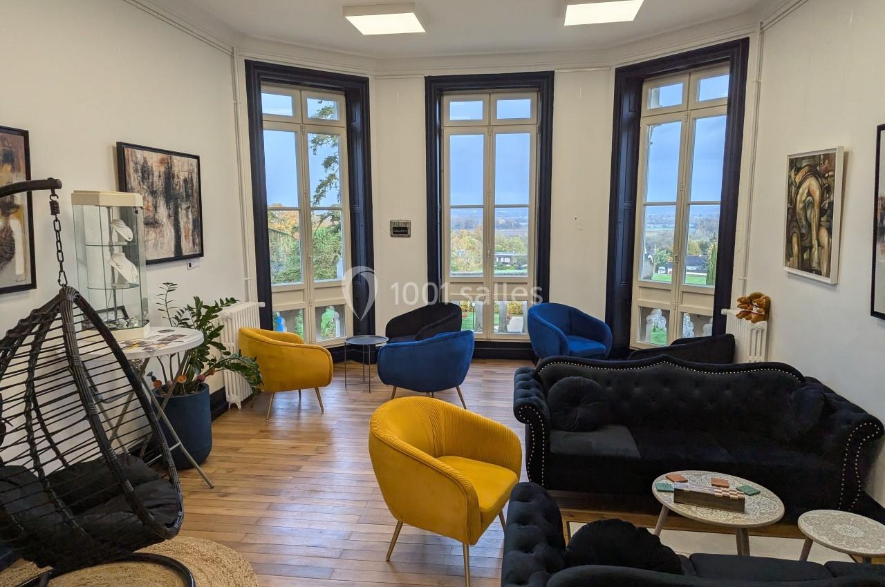 Salon lumineux avec parquet, fauteuils colorés, canapé noir, grandes fenêtres et vue sur un paysage extérieur.