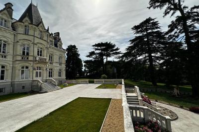 Miniature Terrasse Façade d'un château avec terrasse en pierre, pelouses aménagées et arbres imposants sous un ciel partiellement nuageux.