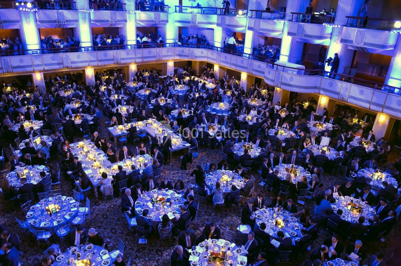 Salle de réception bondée avec des tables rondes dressées, éclairage bleu et jaune, et invités en tenue formelle.