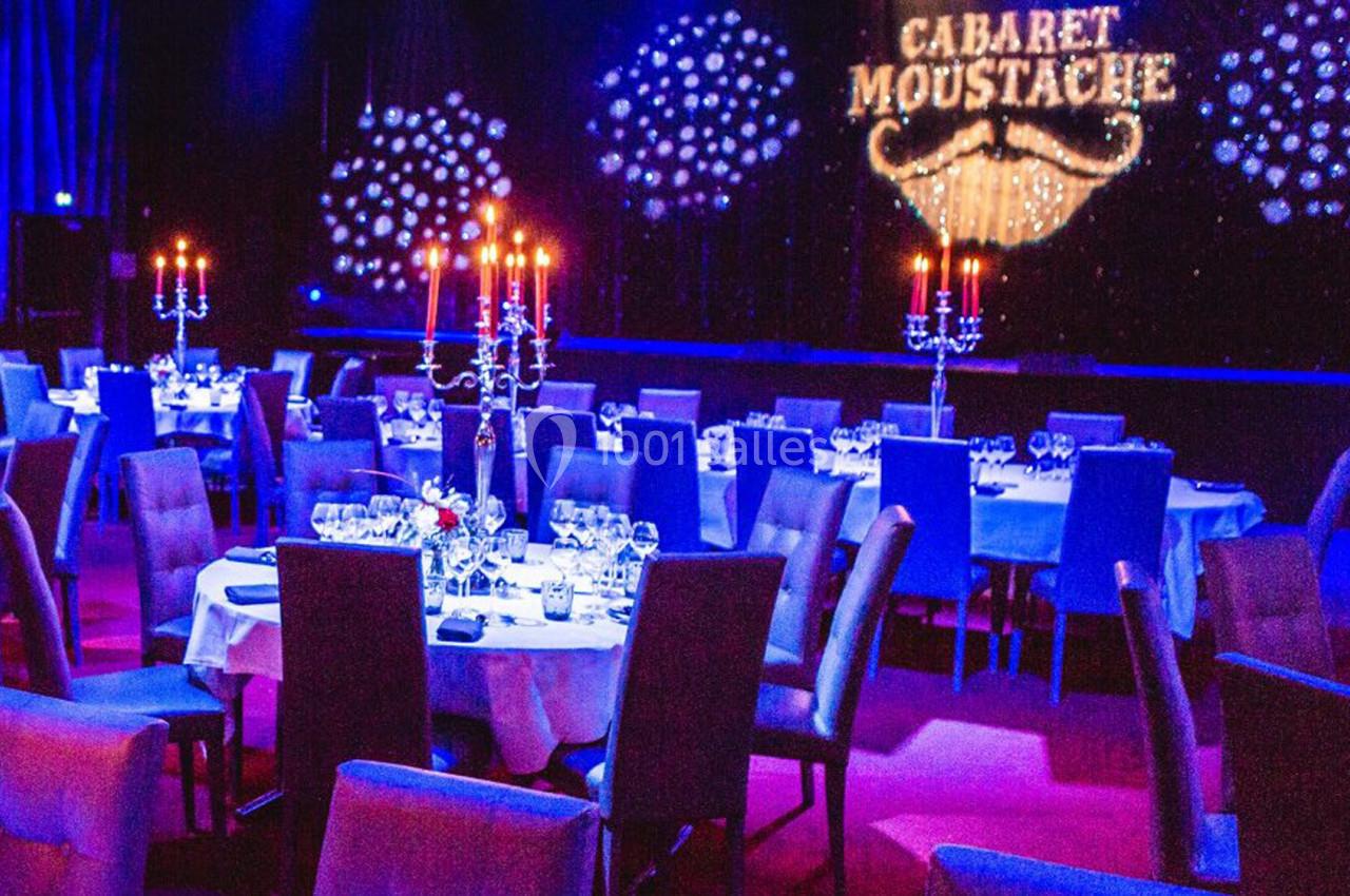 Salle de cabaret avec tables dressées, chandeliers et enseigne lumineuse ’Cabaret Moustache’ en arrière-plan.