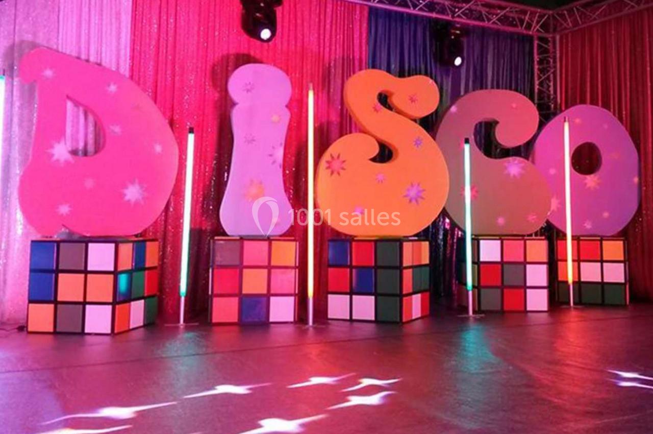 Décor coloré avec des lettres formant ’DISCO’ posées sur des cubes lumineux, entourées de rideaux scintillants.