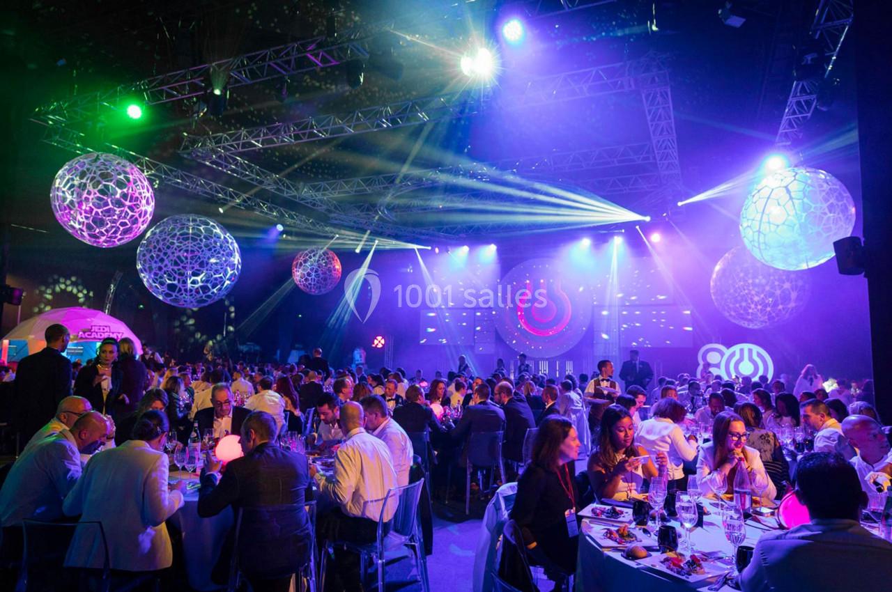 Salle de réception illuminée avec des invités assis à des tables rondes, décorations suspendues et éclairages colorés.