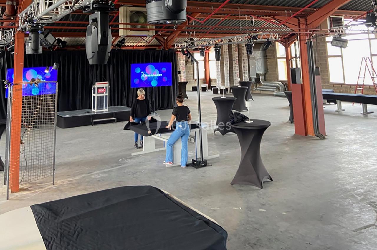 Deux personnes préparent une salle industrielle avec des tables hautes et des écrans pour un événement.