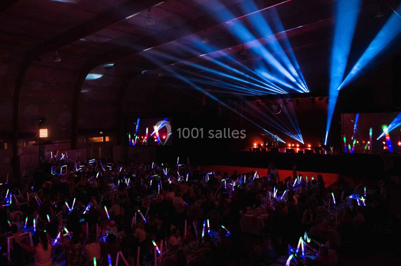Salle sombre avec un public tenant des bâtons lumineux colorés, éclairée par des projecteurs et des faisceaux lumineux.