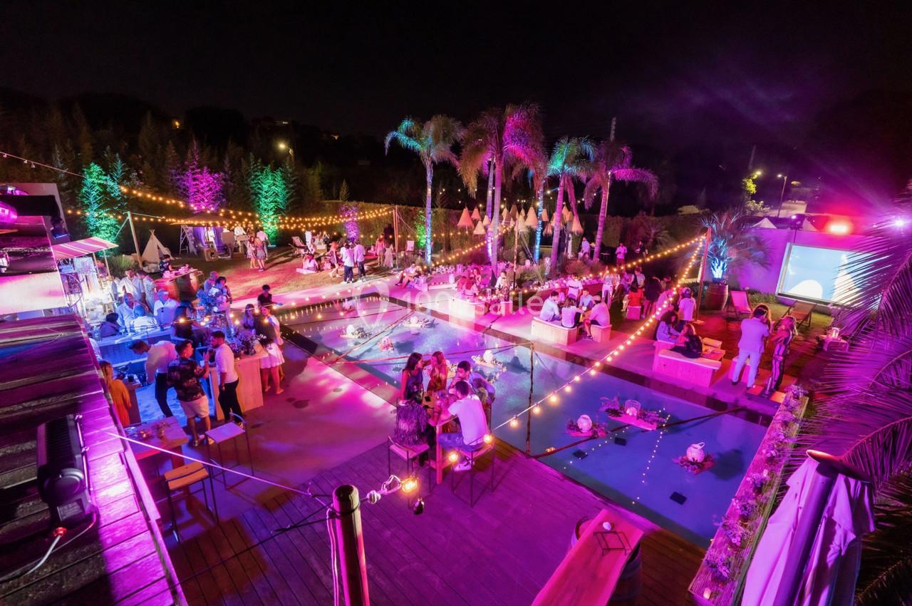 Soirée festive en extérieur autour d'une piscine éclairée, avec invités, décorations lumineuses et palmiers.