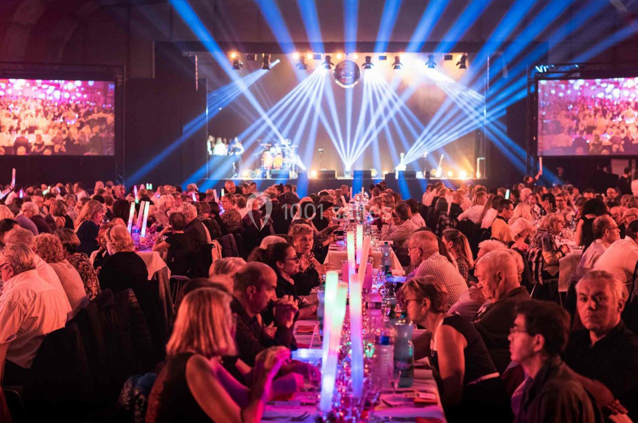 Salle comble avec des tables alignées, des convives attablés et une scène illuminée de projecteurs en arrière-plan.