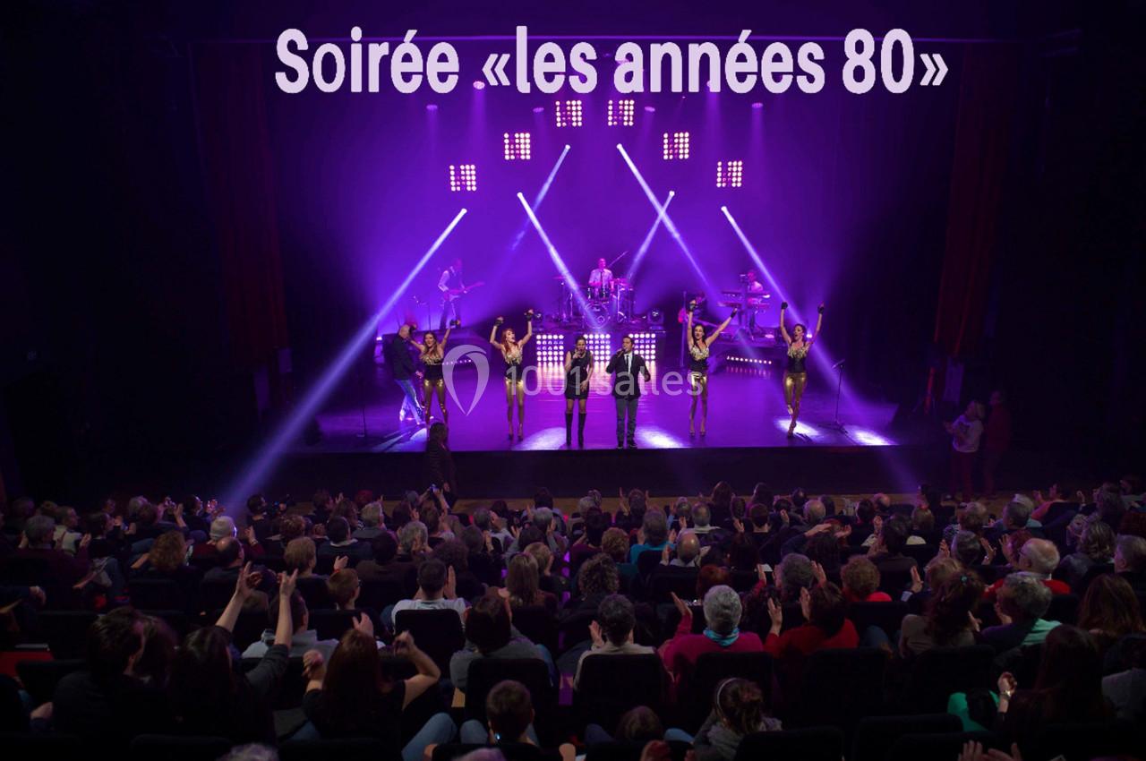 Spectacle sur scène avec musiciens et danseurs, éclairage violet, devant un public assis dans une salle comble.