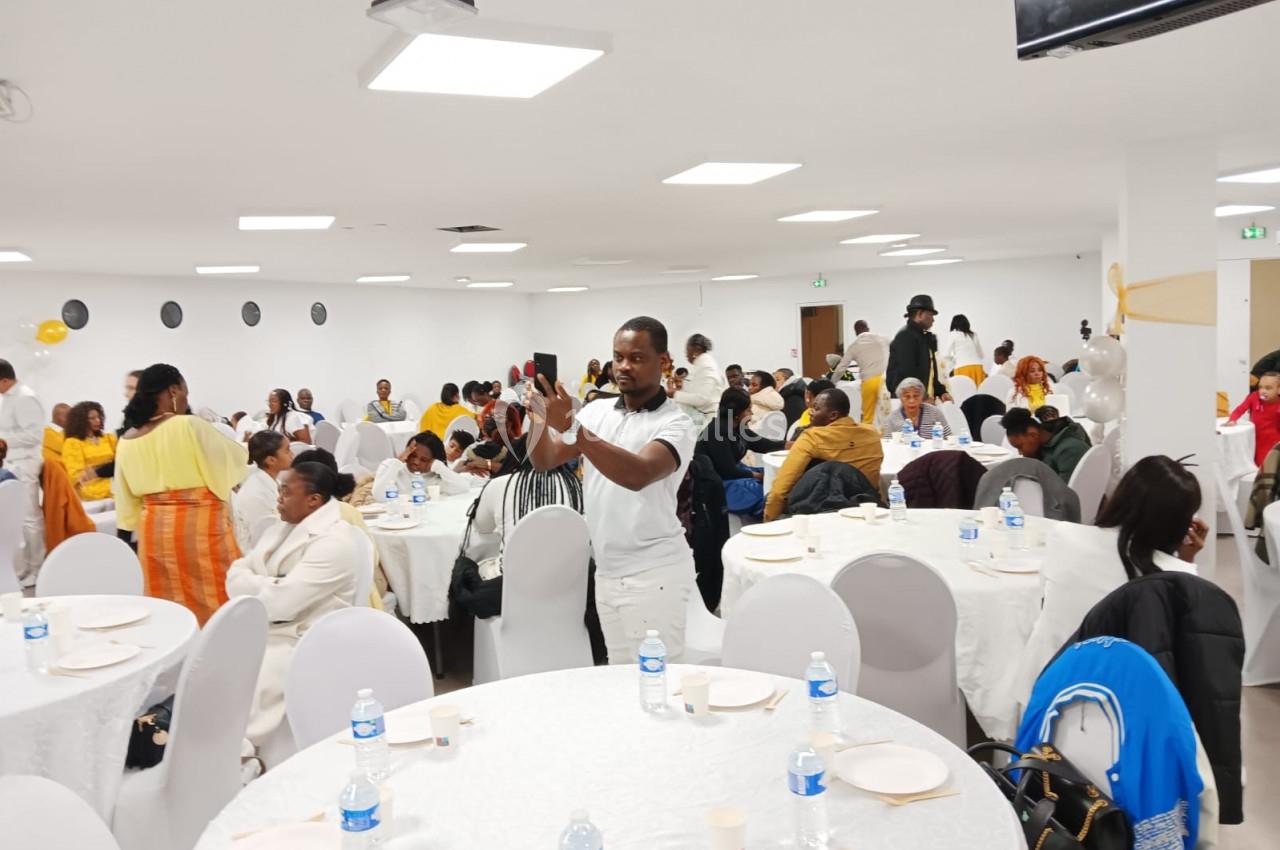 Des personnes assises autour de tables rondes dans une salle lumineuse, avec un homme prenant une photo au centre.