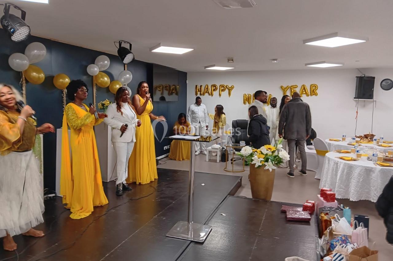 Groupe de personnes en tenue festive, certaines chantant sur une scène, décorations et tables de fête en arrière-plan.