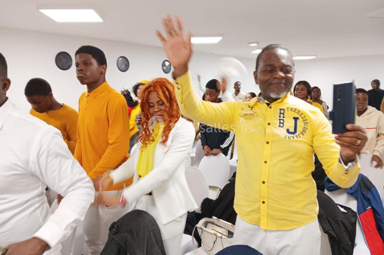 Des personnes debout dans une salle lumineuse, certaines souriantes et saluant, vêtues de blanc et de jaune.