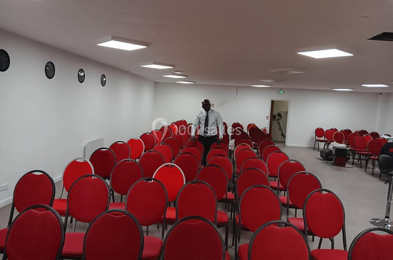 Un homme marche entre des rangées de chaises rouges alignées dans une salle blanche éclairée.