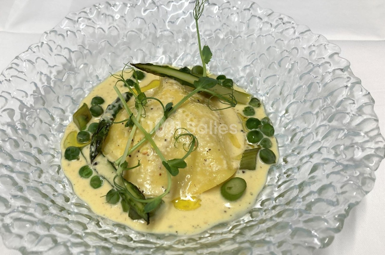 Ravioles servies dans une assiette en verre, accompagnées de sauce crémeuse, petits pois et pousses vertes.