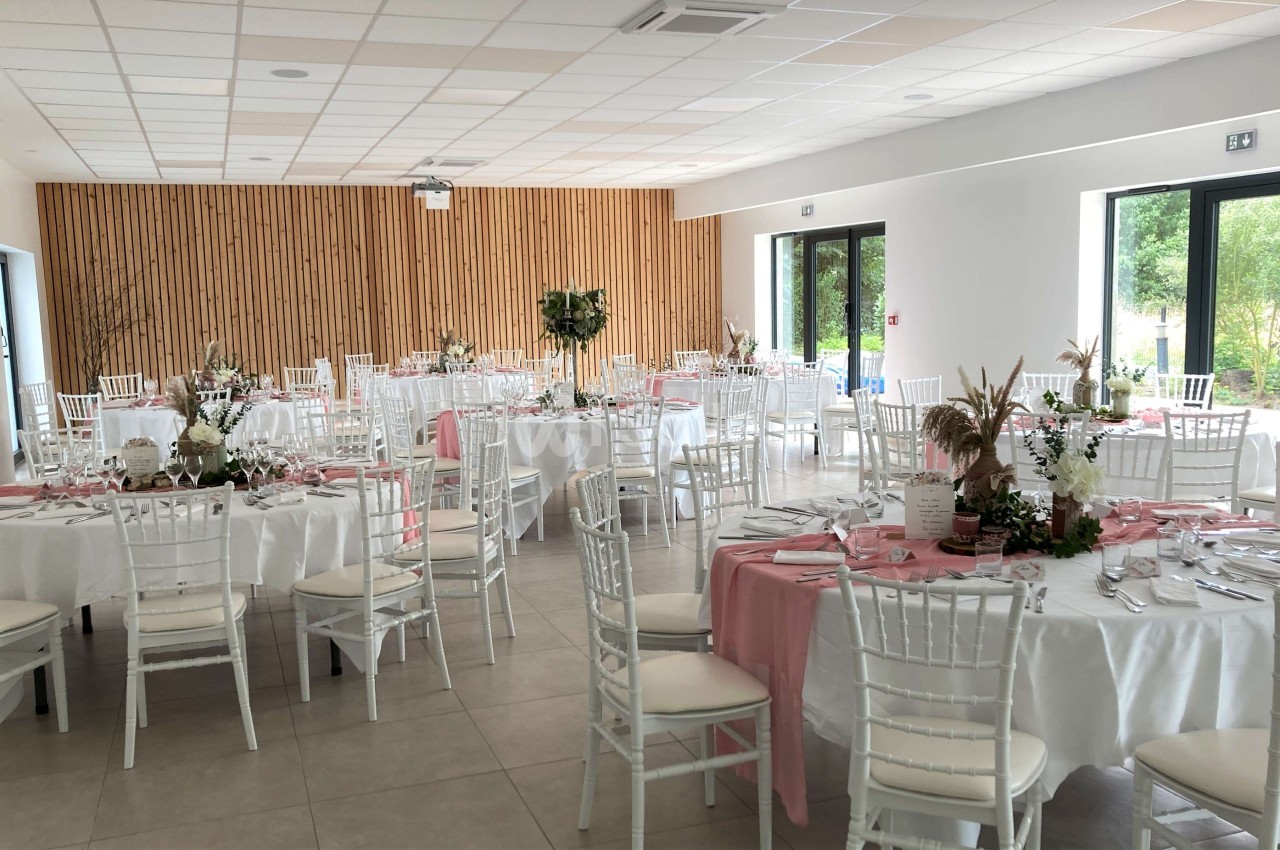 Salle de réception lumineuse avec tables rondes dressées, nappes blanches, décorations florales et chaises blanches.