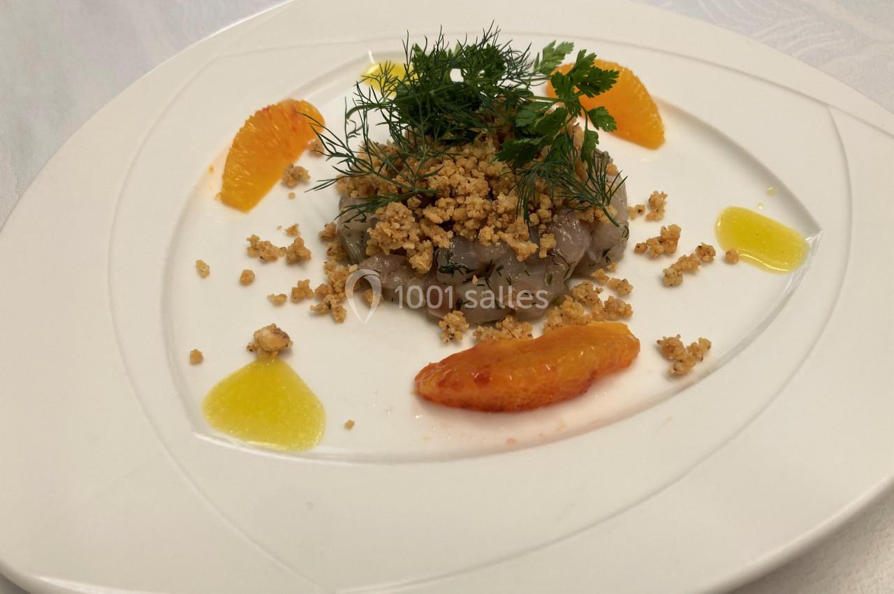 Tartare de poisson présenté avec des segments d'orange, crumble, herbes fraîches et sauce jaune sur une assiette blanche.