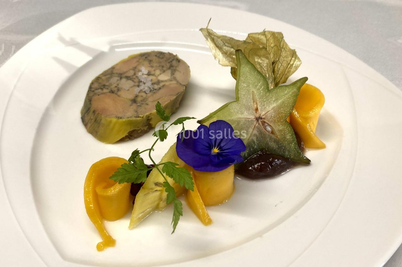 Assiette gastronomique avec terrine, légumes colorés, physalis, carambole et fleur comestible sur une table blanche.