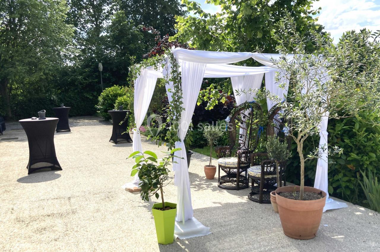 Tonnelle blanche décorée de plantes, entourée de verdure, avec des fauteuils en osier sur une terrasse ensoleillée.