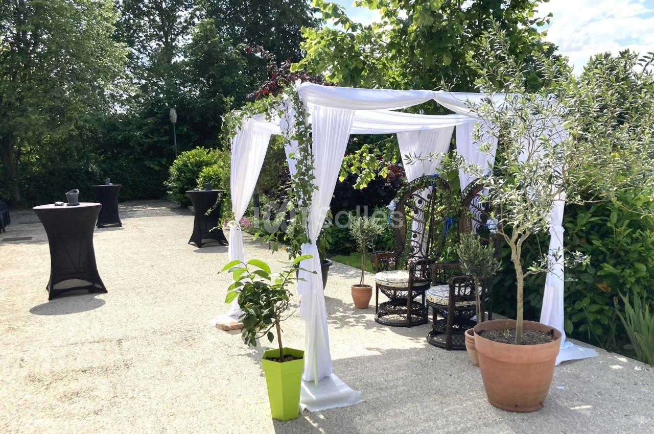 Tonnelle blanche décorée de plantes, entourée de verdure, avec des fauteuils en osier sur une terrasse ensoleillée.