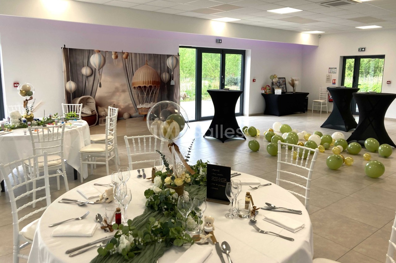 Salle décorée pour un événement avec tables dressées, ballons verts au sol et fond photo avec montgolfière.