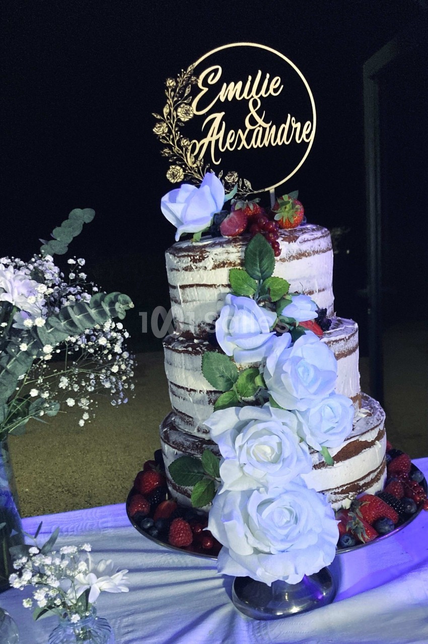 Gâteau de mariage à trois étages décoré de roses blanches, fruits rouges et un topper personnalisé.