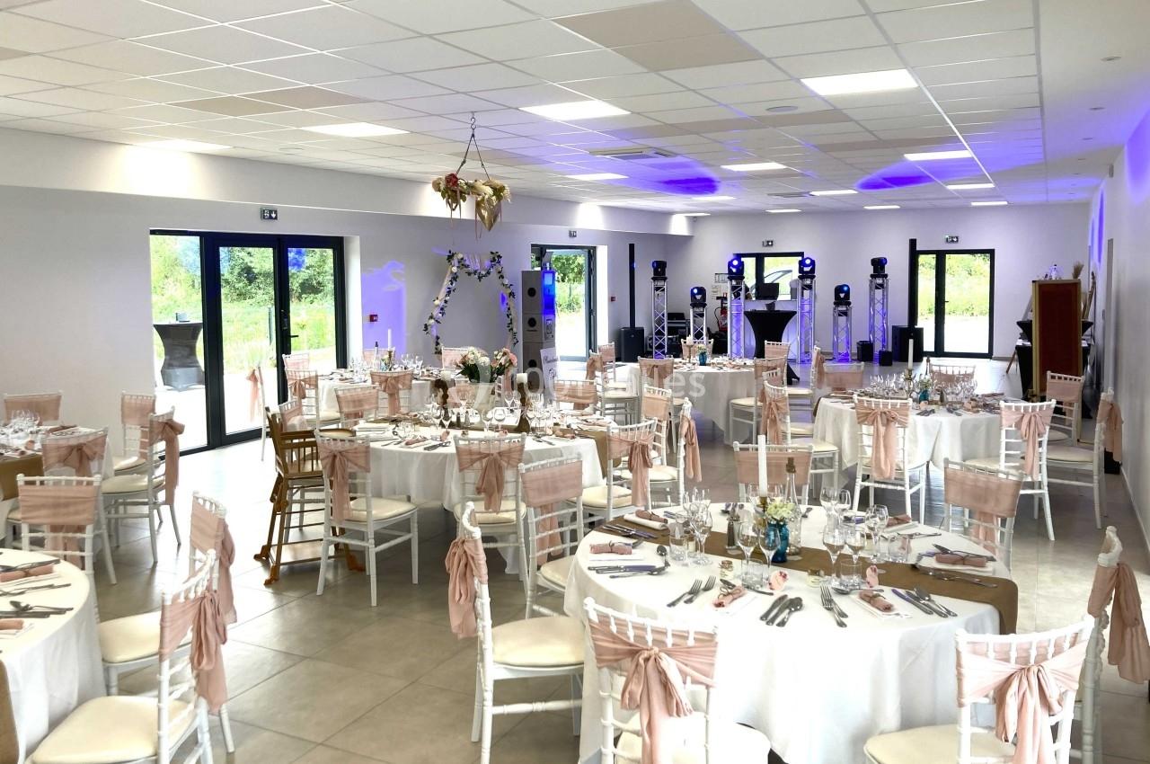 Salle de réception décorée avec des tables rondes dressées, chaises blanches ornées de nœuds roses et éclairage d'ambiance.