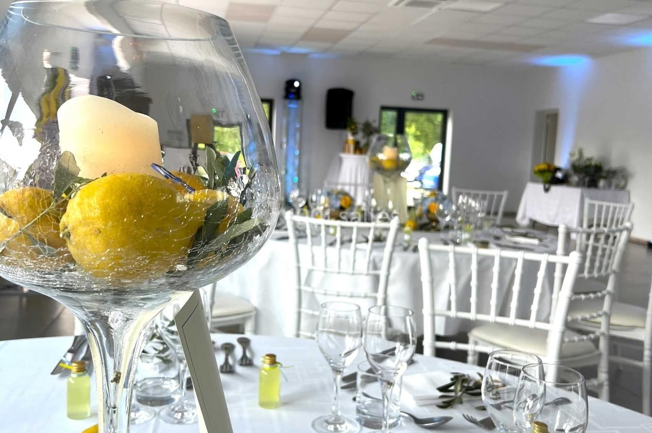 Salle de réception décorée avec des tables dressées, un centre de table avec bougie, citrons et fleurs au premier plan.