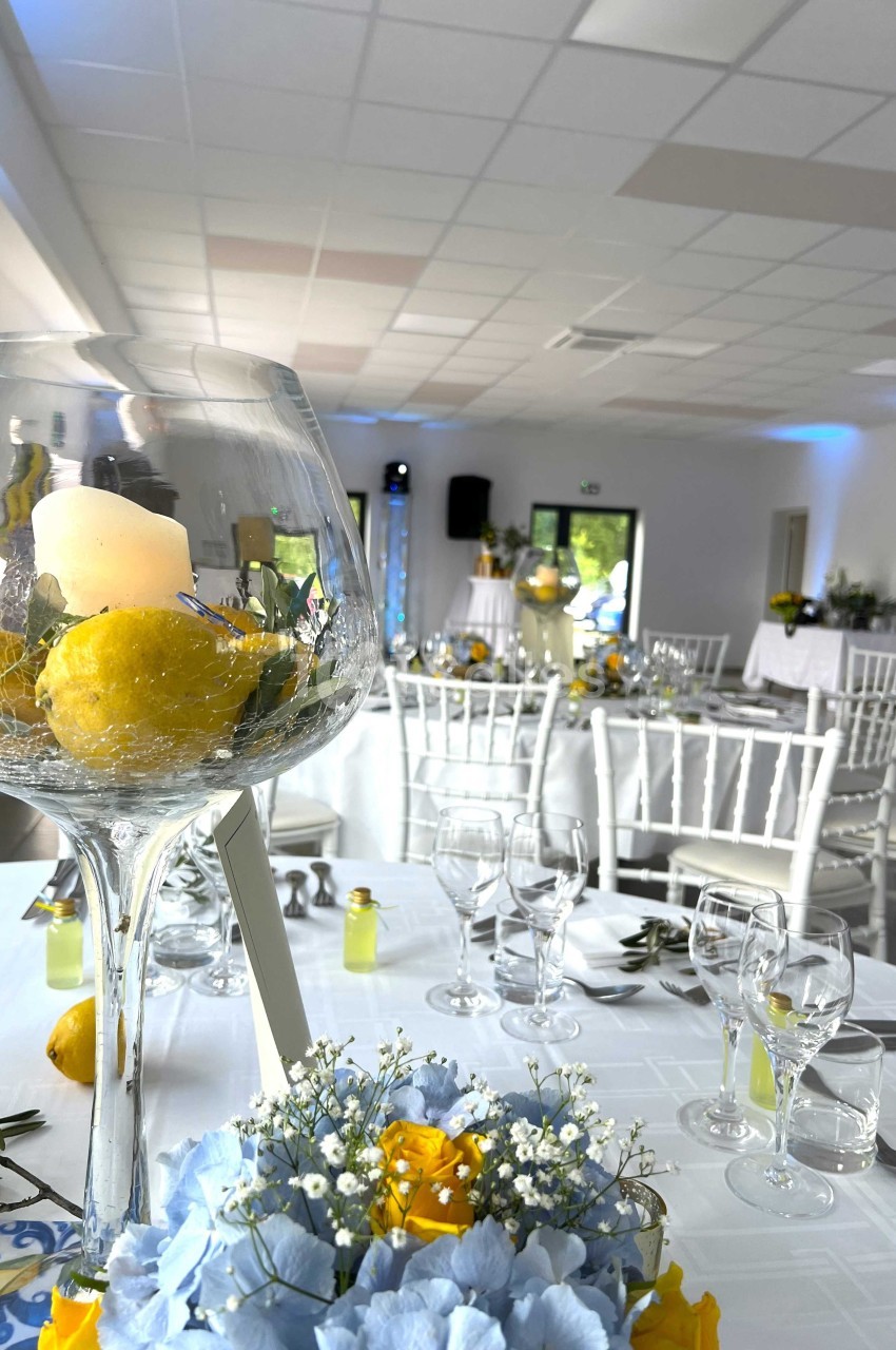 Salle de réception décorée avec des tables dressées, un centre de table avec bougie, citrons et fleurs au premier plan.