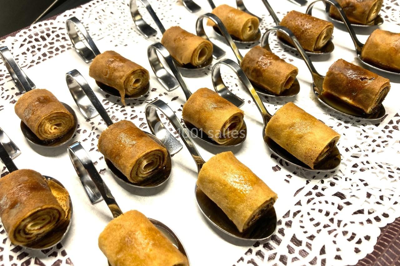 Petits roulés dorés disposés sur des cuillères en métal, présentés sur une nappe en dentelle blanche.