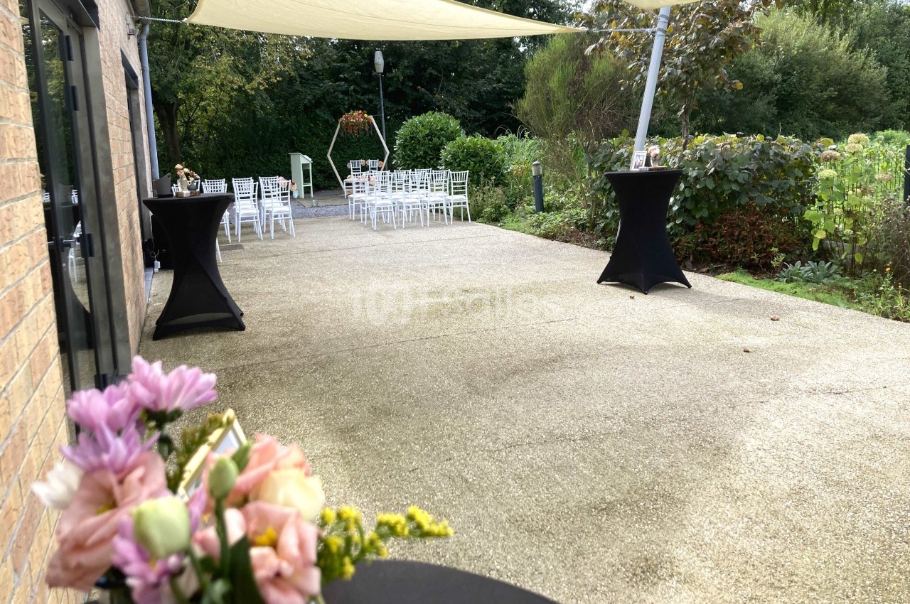 Terrasse aménagée pour un événement avec chaises blanches, tables hautes et décorations florales, entourée de verdure.