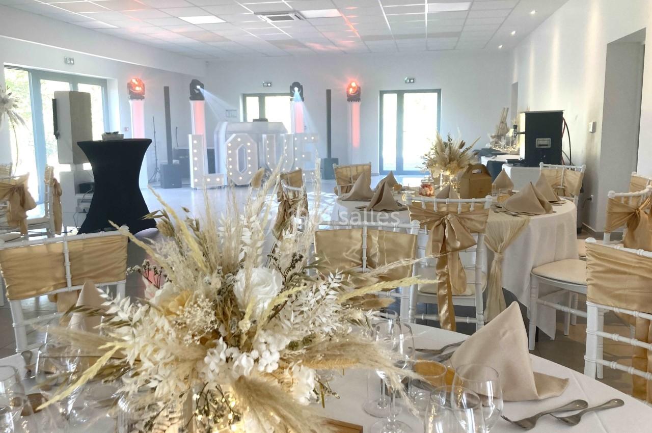 Salle de réception décorée pour un mariage, avec tables dressées, chaises ornées de rubans dorés et un grand panneau ’LOVE’.