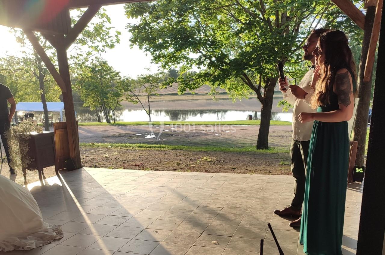Un couple debout sous une pergola regarde vers un lac entouré d'arbres au coucher du soleil.