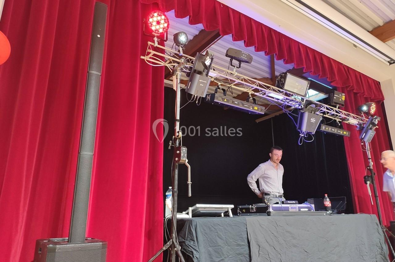 Un DJ se tient derrière une table équipée de matériel audio et d'éclairage sur une scène avec rideaux rouges.