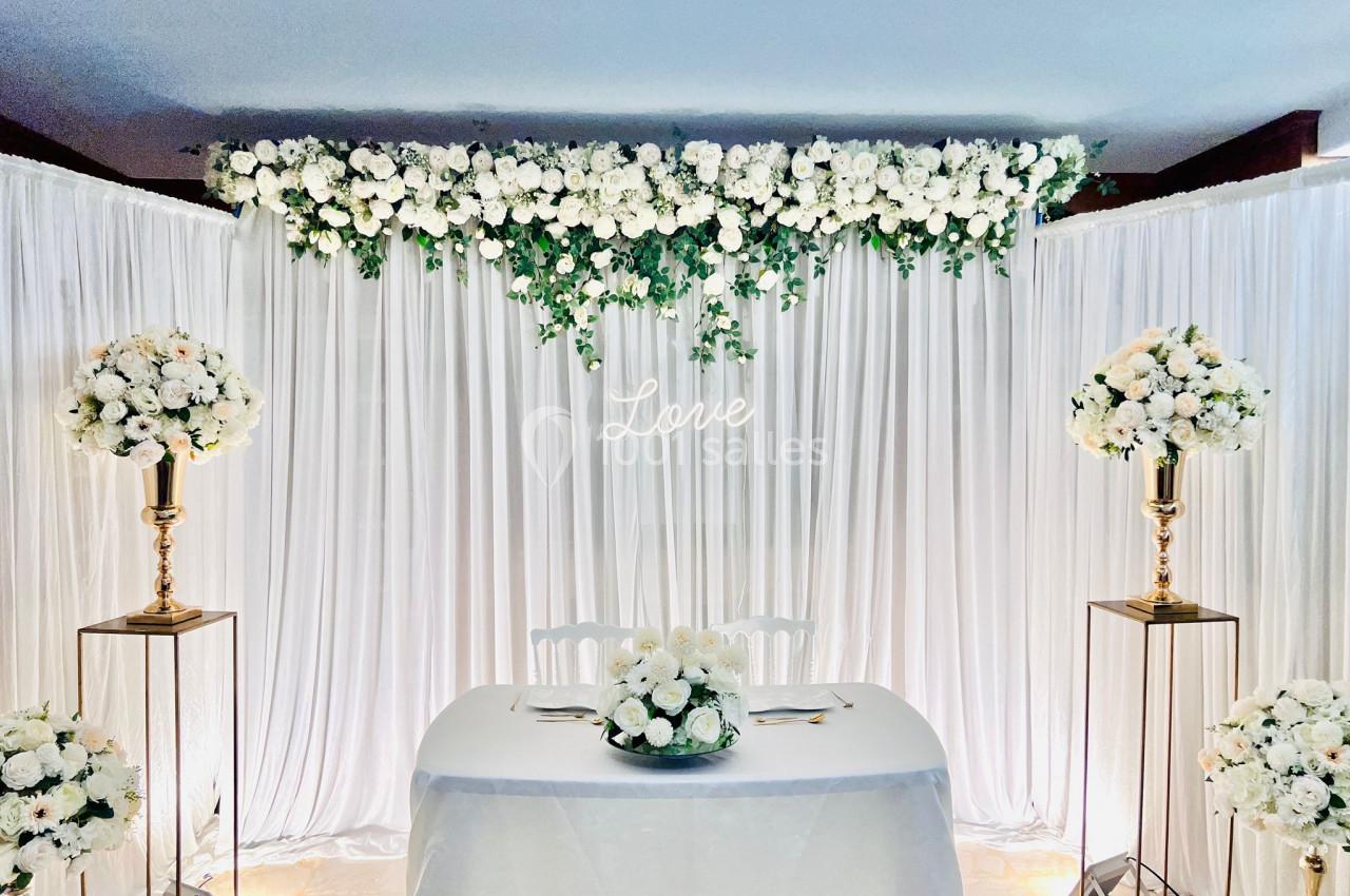 Décoration de mariage avec rideaux blancs, guirlande de fleurs blanches et vertes, et table ornée d'un bouquet.