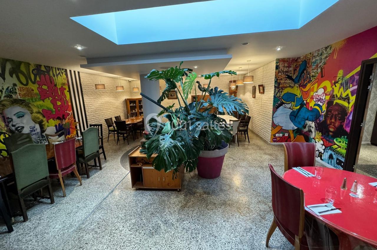 Salle de restaurant moderne avec fresques murales colorées, mobilier varié et grande plante en pot au centre.
