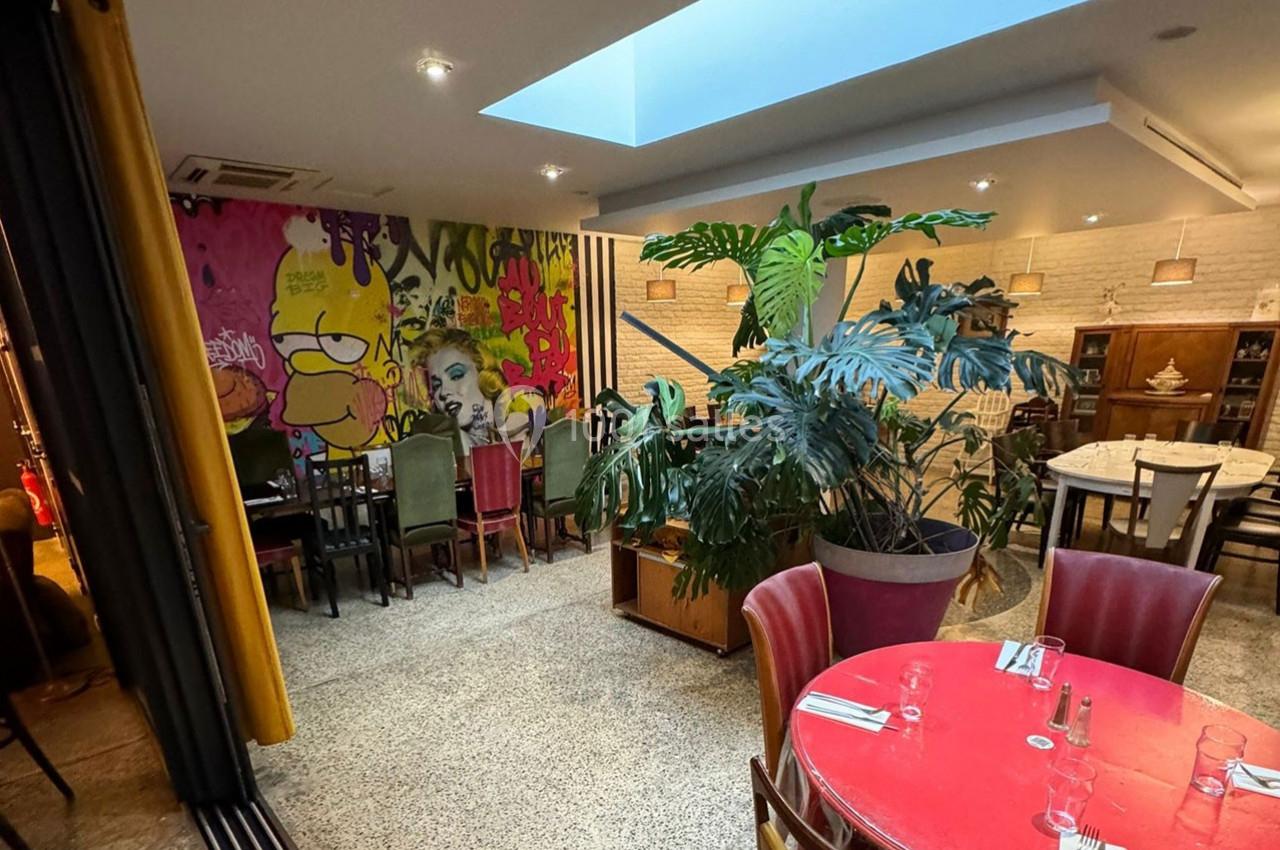 Salle de restaurant avec tables colorées, plantes, fresque murale artistique et éclairage naturel par une verrière.