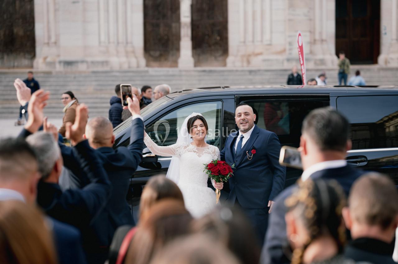 Un couple de mariés souriant pose devant une voiture noire, saluant une foule rassemblée devant un bâtiment.