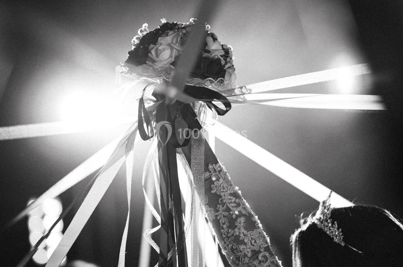 Une mariée lève un bouquet entouré de rubans lors d'une célébration, éclairée par des lumières en arrière-plan.