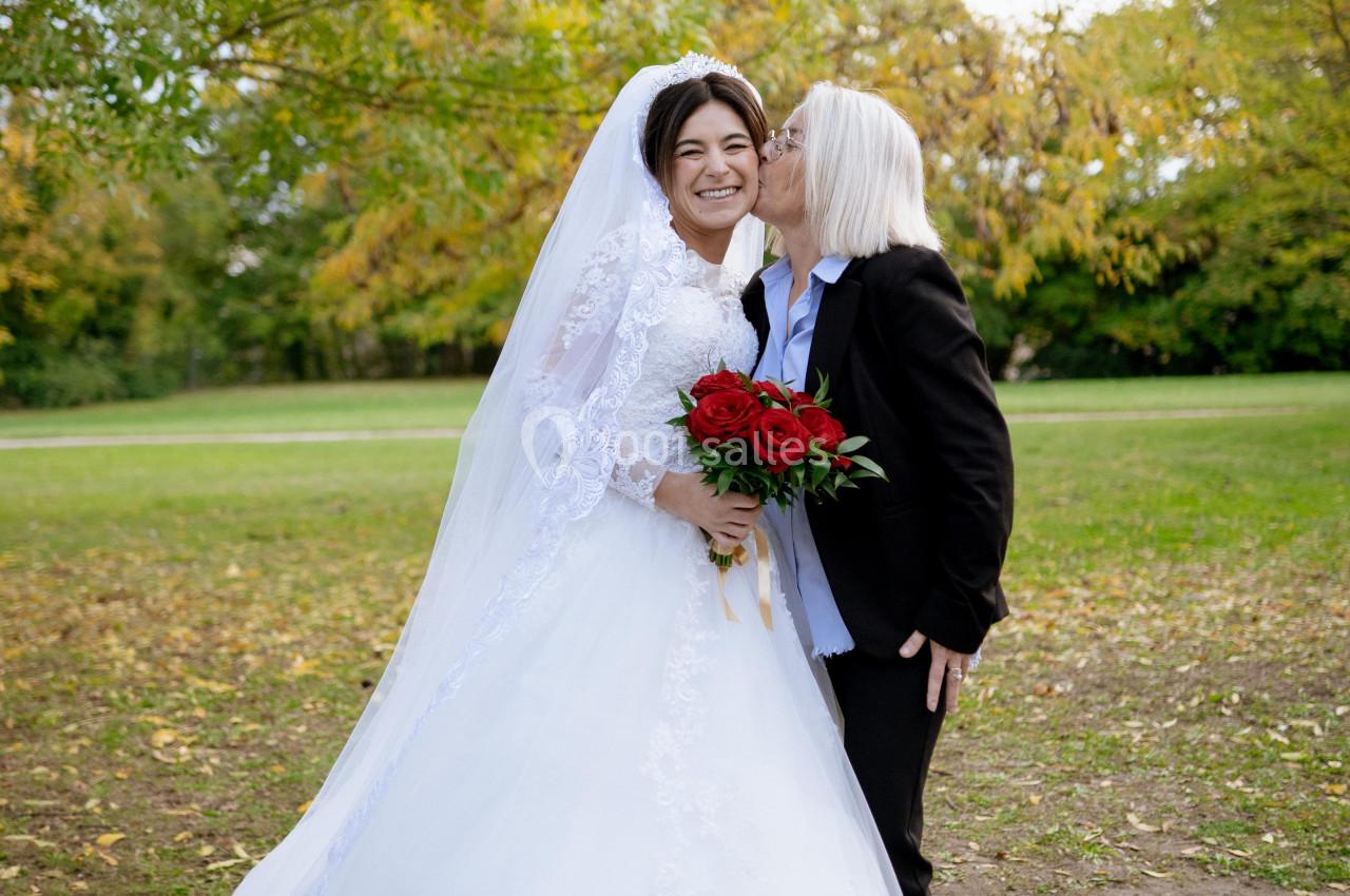 Une mariée souriante en robe blanche est embrassée sur la joue par une personne en costume noir dans un parc.
