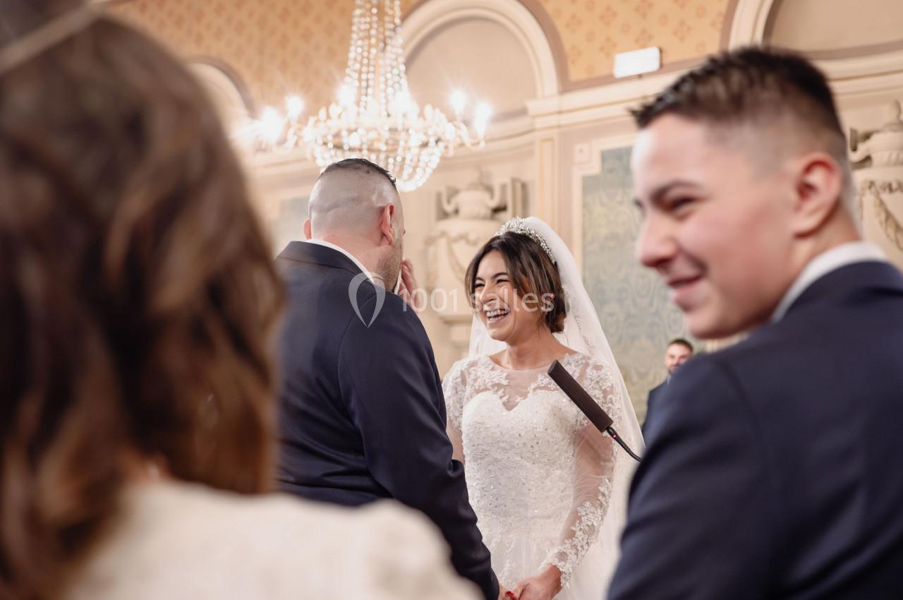 Un couple échange ses vœux de mariage dans une salle ornée de fresques et d'un lustre imposant.