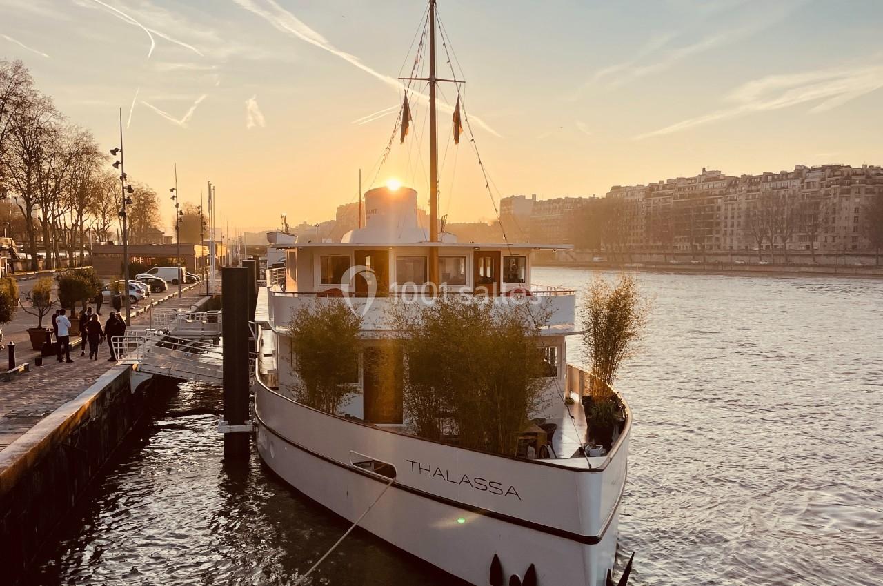 Bateau amarré sur une rivière au coucher du soleil, avec des arbres et des bâtiments en arrière-plan.