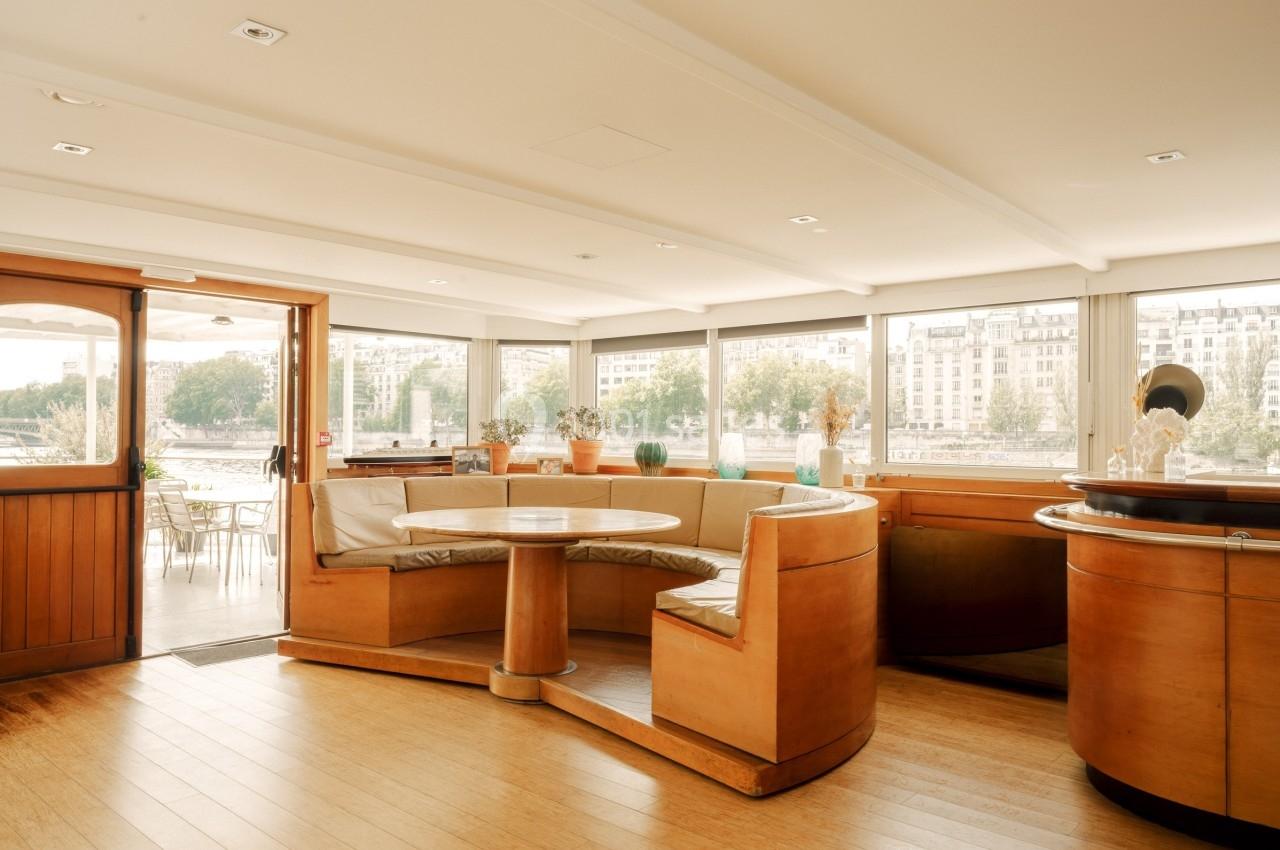 Intérieur lumineux d'une péniche avec banquettes en bois, table ronde et vue sur des bâtiments au bord de l'eau.
