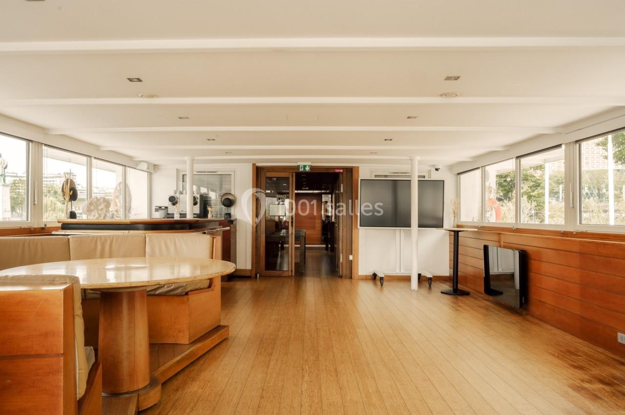 Intérieur spacieux et lumineux d'un bateau avec parquet, mobilier en bois et grandes fenêtres offrant une vue extérieure.